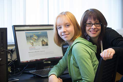 Amy Siebert-McKenzie and Marisa Brake pose in their computer lab with the conference website pulled up on screen