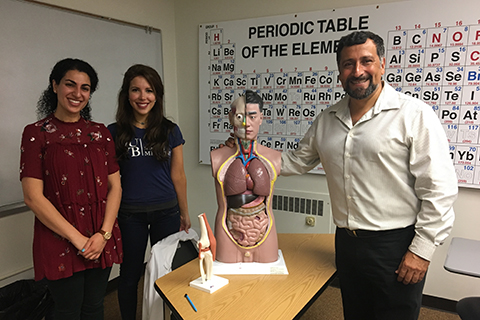 professor standing in front of a periodic table of the elements chart, holding a model of a human torso, with two students on his right