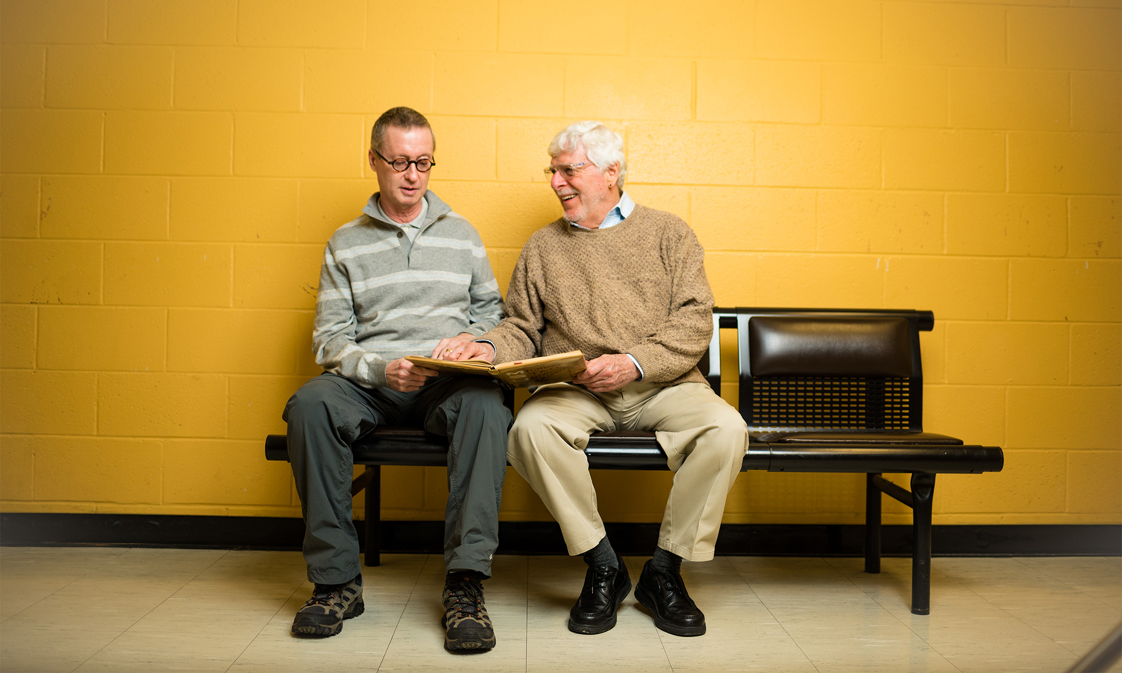 Oakland University alumni Bruce Voss and Al Monetta sit in front of a gold wall on campus