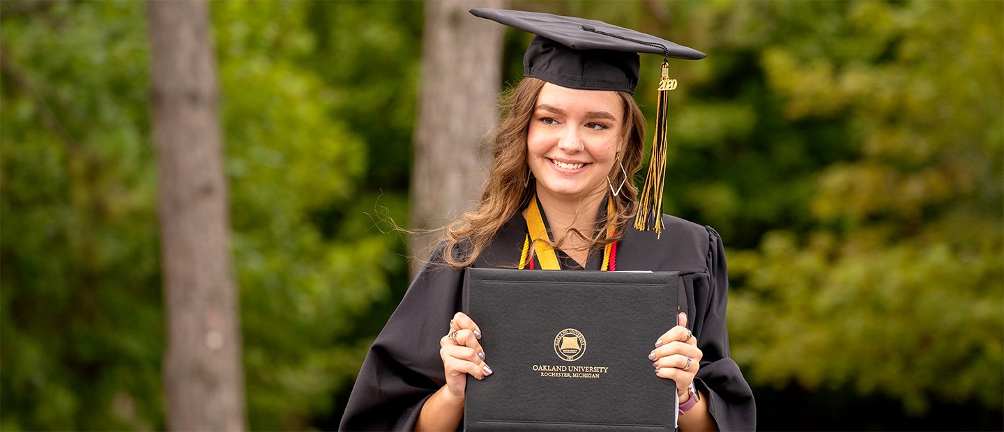 A female Oakland University graduate, wearing a cap and gown, holding up a diploma.