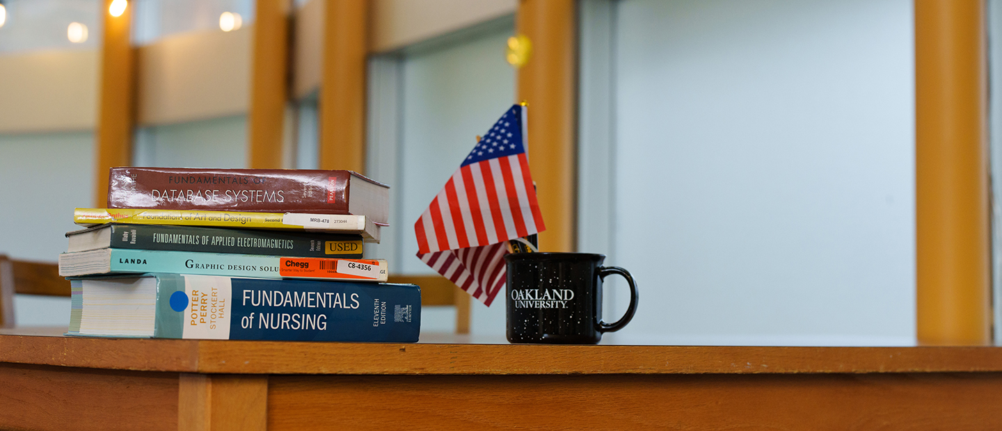 A pile of text books sits on a desk along with an Oakland University coffee mug that contains small American flags.