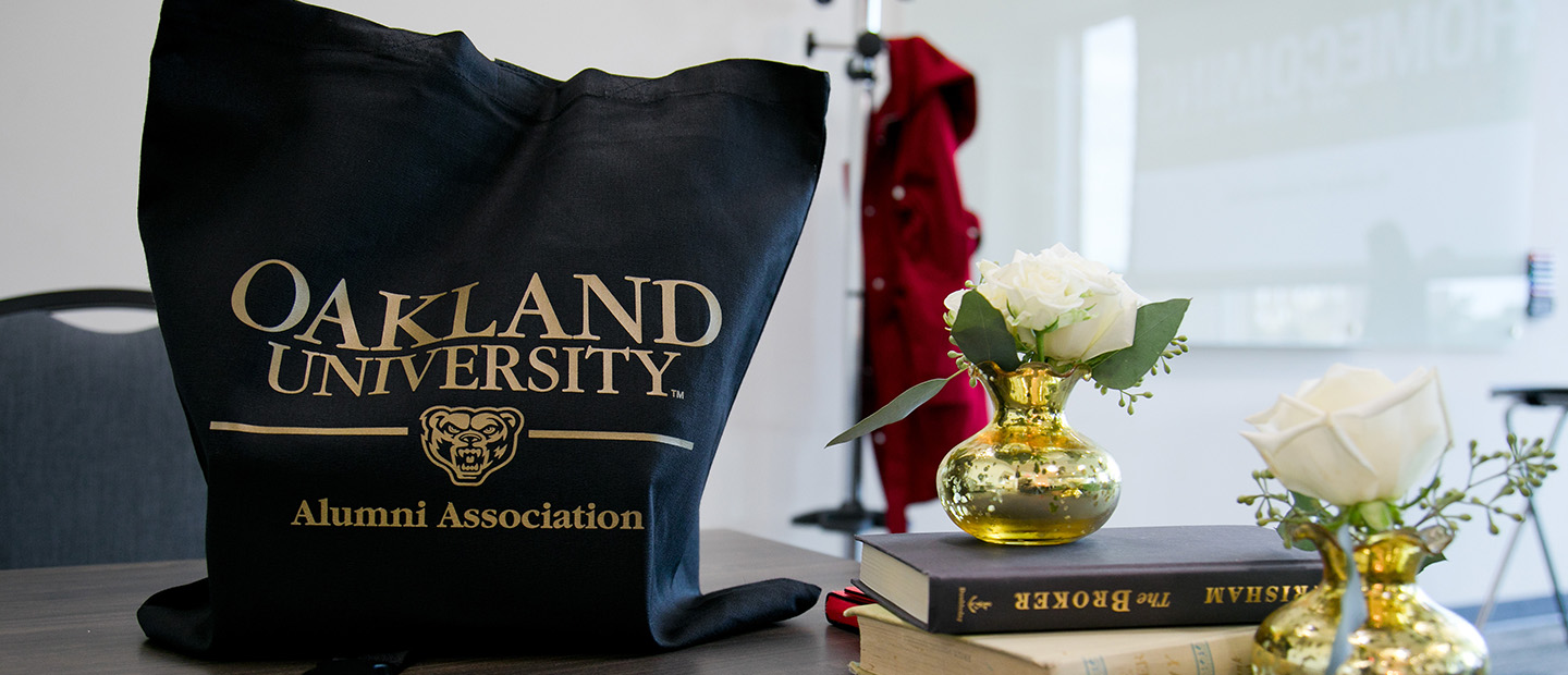 Oakland University Alumni Association bag on a table next to books and flowers in vases.