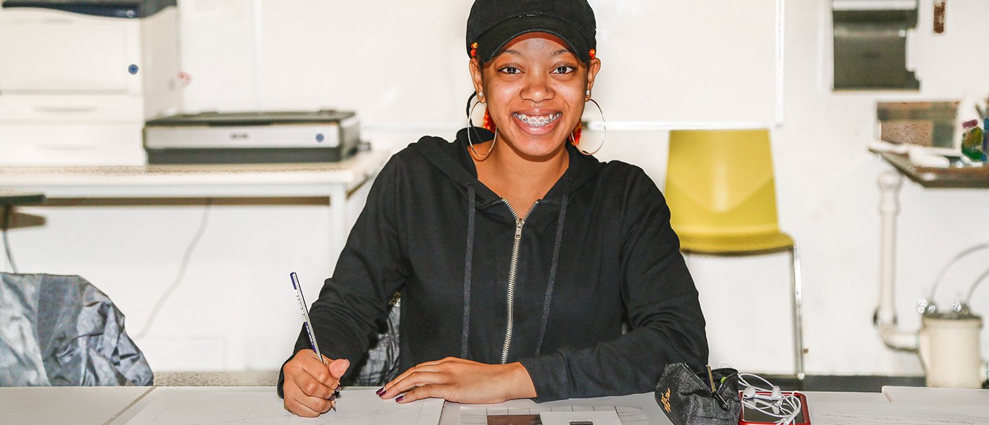 woman seated at a desk with pen and paper, smiling at the camera