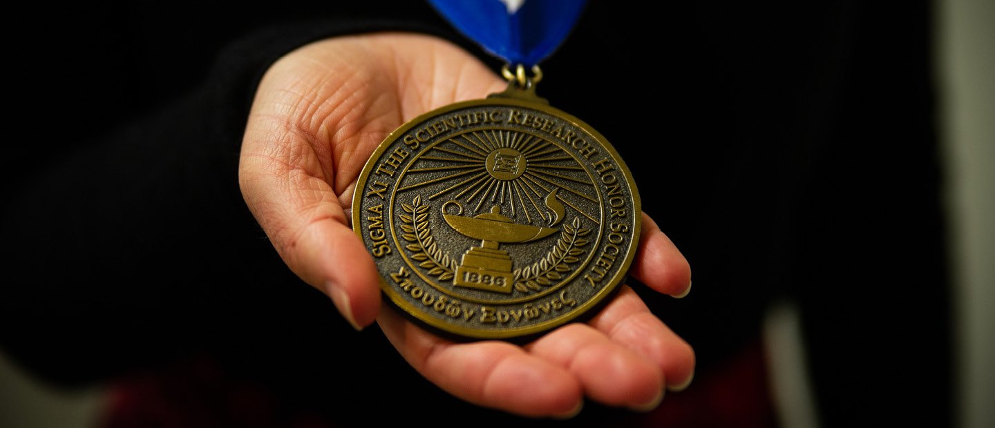 A hand holding a round gold medal. Text on the medal says "Sigma Xi The Scientific Research Honor Society"