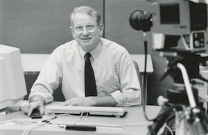 John Tower seated at a desk, being filmed with a video camera.
