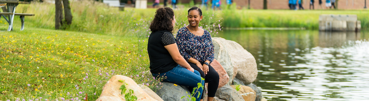 Image of two students sitting by a lake having a conversation. 