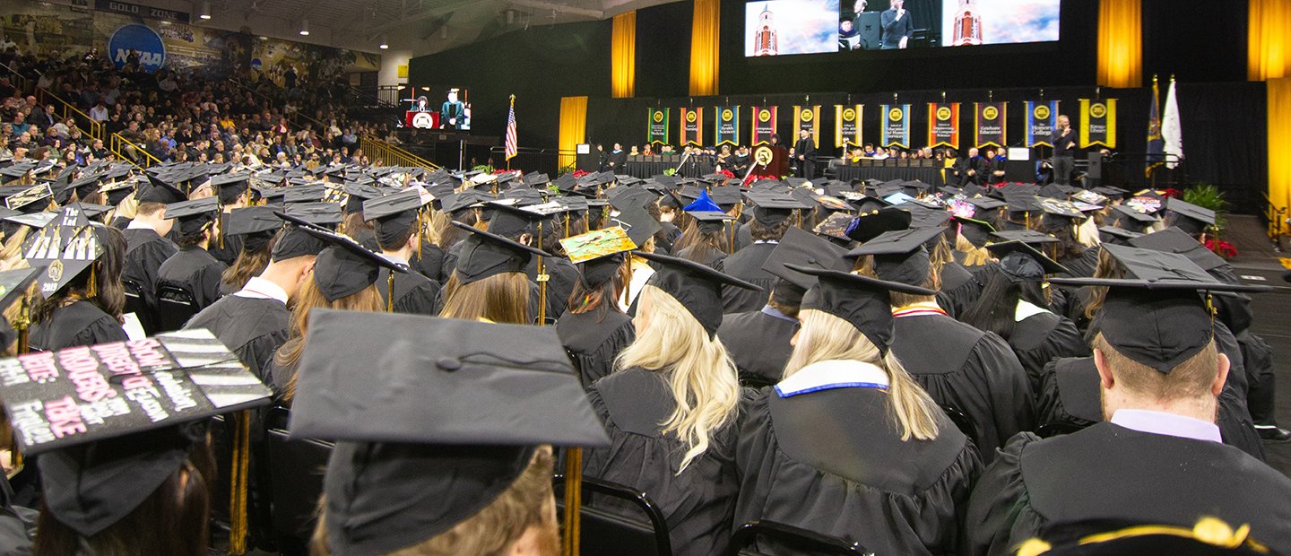 A photo of graduates in caps and gowns, facing a stage.