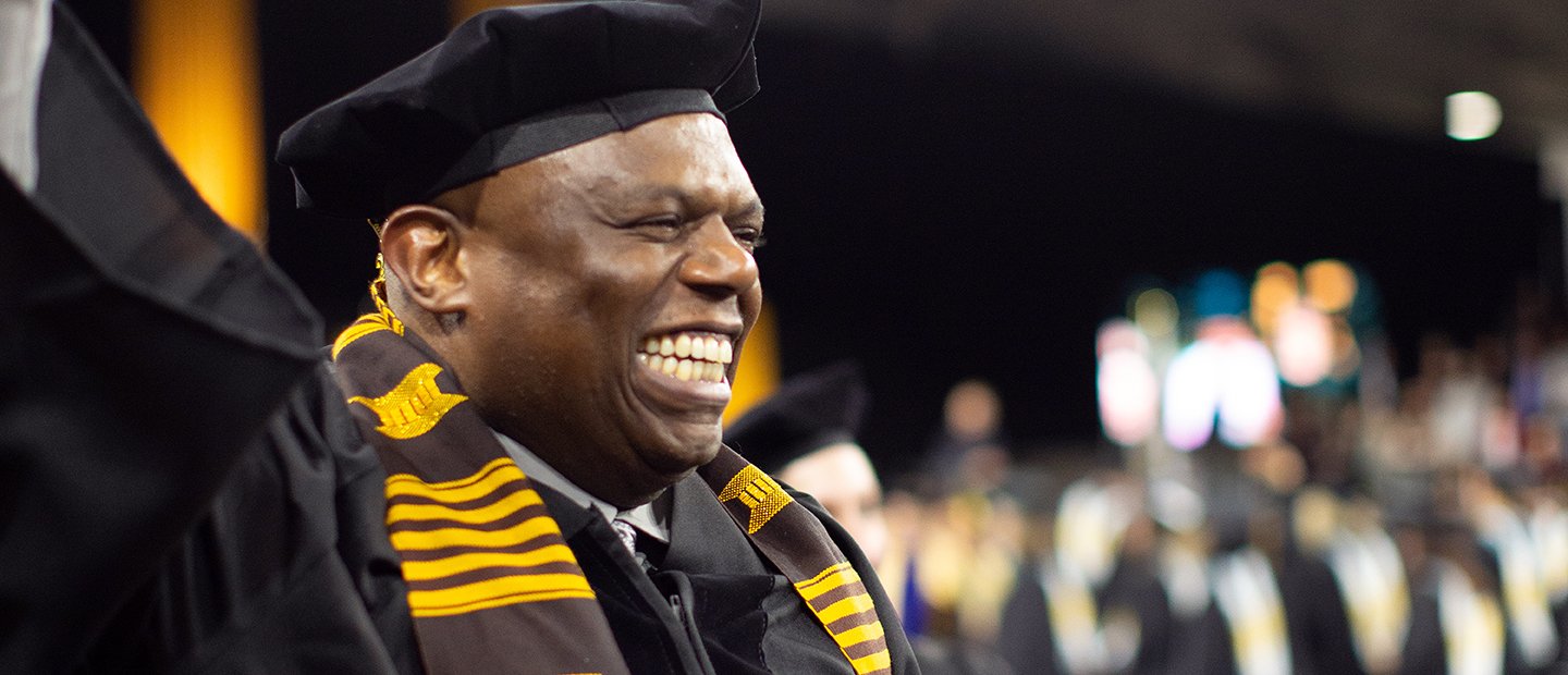 A man in a black cap and gown triumphantly raising his arm and smiling.
