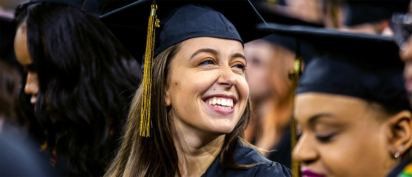 A young woman wearing a cap and gown, smiling, at a graduation ceremony.