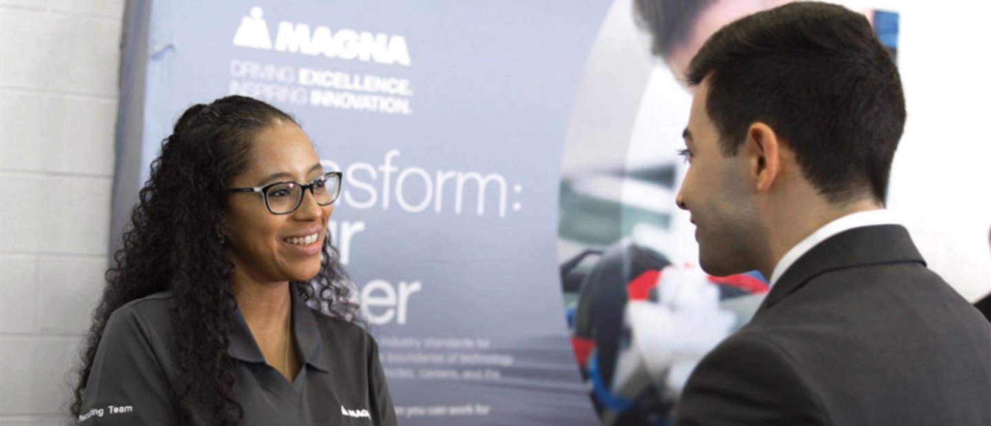 man and woman facing each other, smiling, in front of a sign that says "Magna"