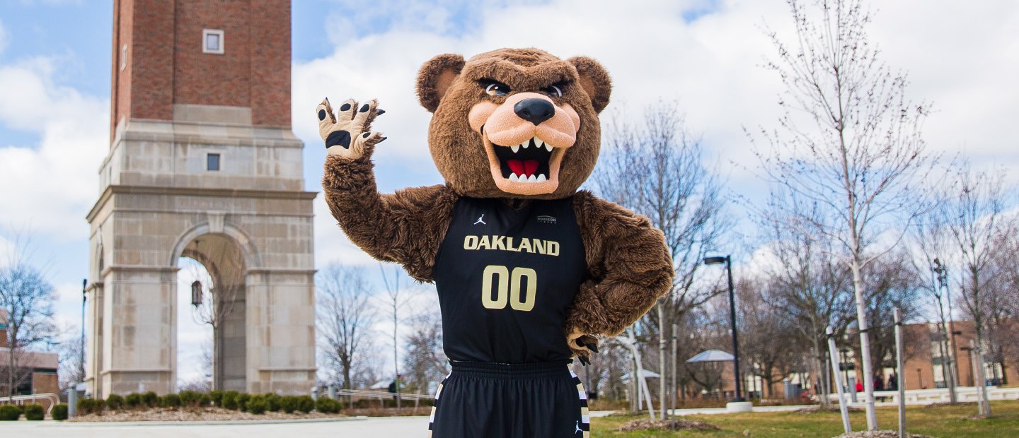The Grizz bear mascot standing outdoors, waving, with Elliott Tower in the background