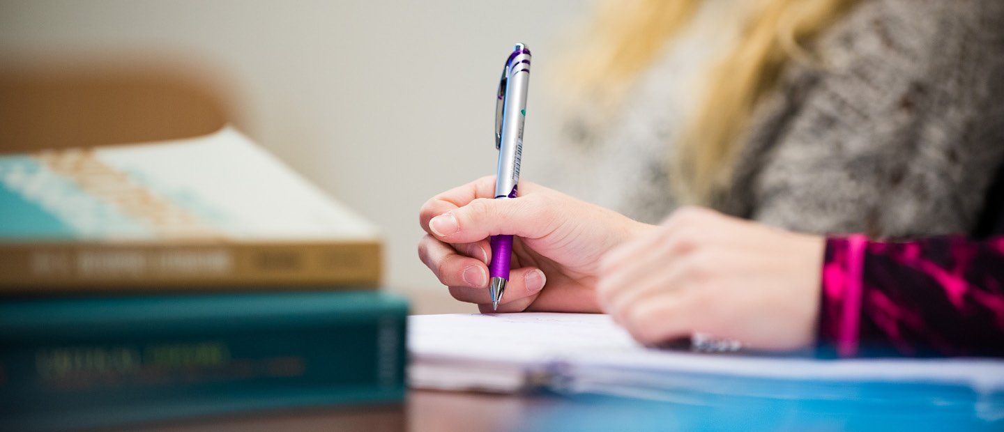 A hand holding a pen over a notebook, next to a stack of books.