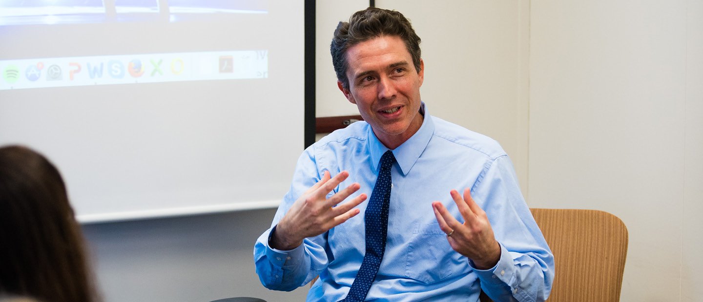 A professor seated in front of a projector screen, taking and gesturing with his hands.