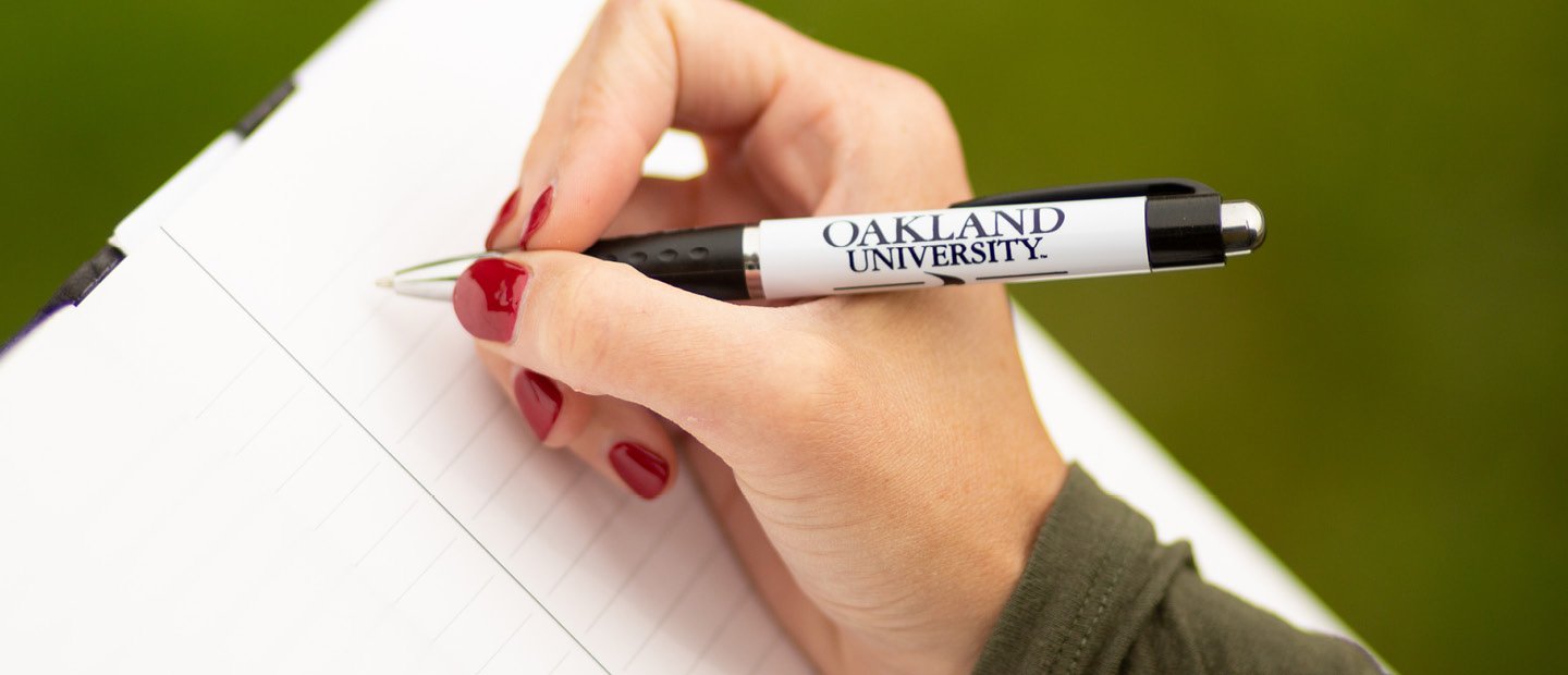 A hand holding an Oakland University pen over a notebook.