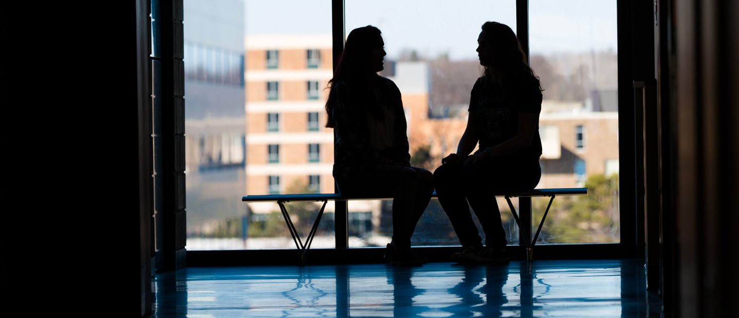 Silhouettes of two people seated on a bench in front of a window with O U campus in the background
