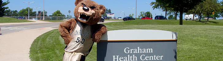 The Grizz bear mascot leaning on a sign outdoors that says Graham Health Center.