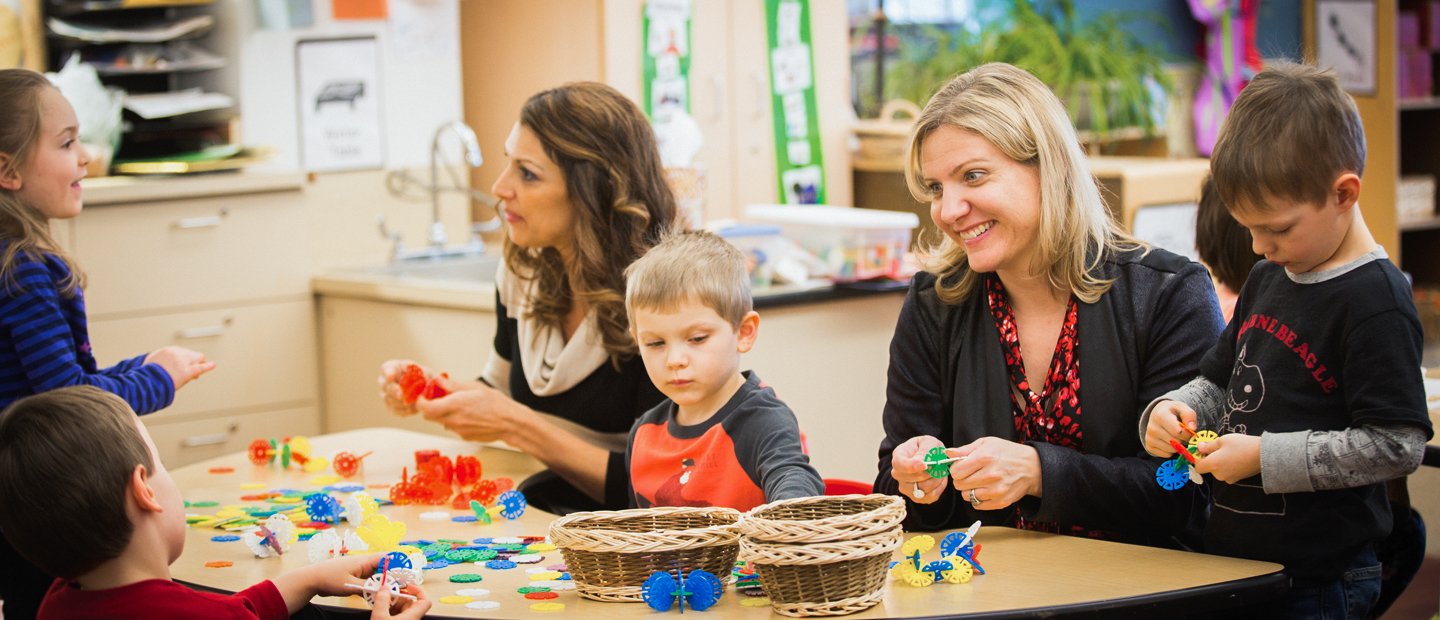 Two female teachers with four young students playing with colorful toys in a classroom.