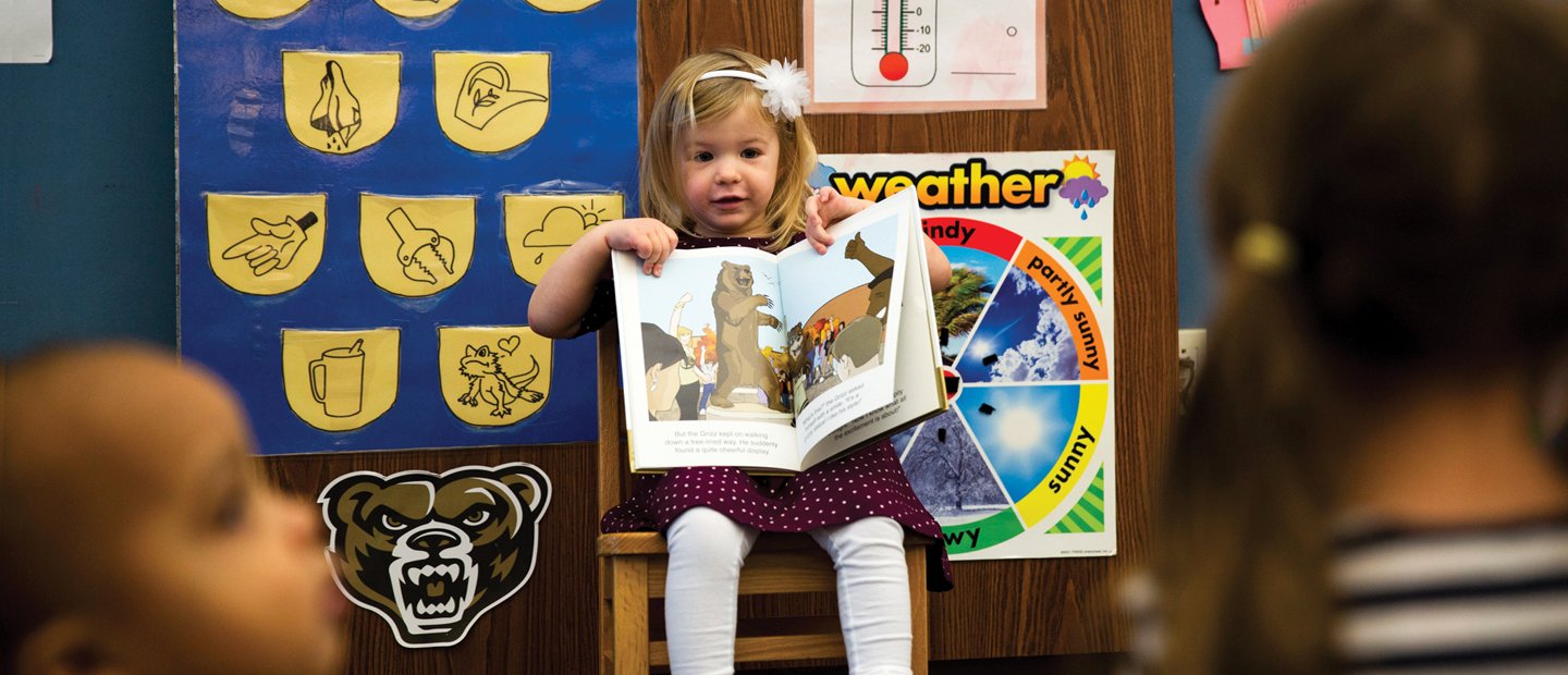 Young girl sitting at the front of a classroom, showing the other students a book.