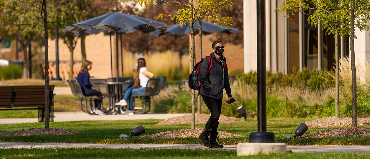 A student wearing a face mask, walking on Oakland University's campus. Two students in masks are sitting at a table behind her.
