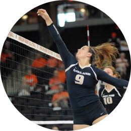 woman in an Oakland jersey jumping with her arm extended over a volleyball net