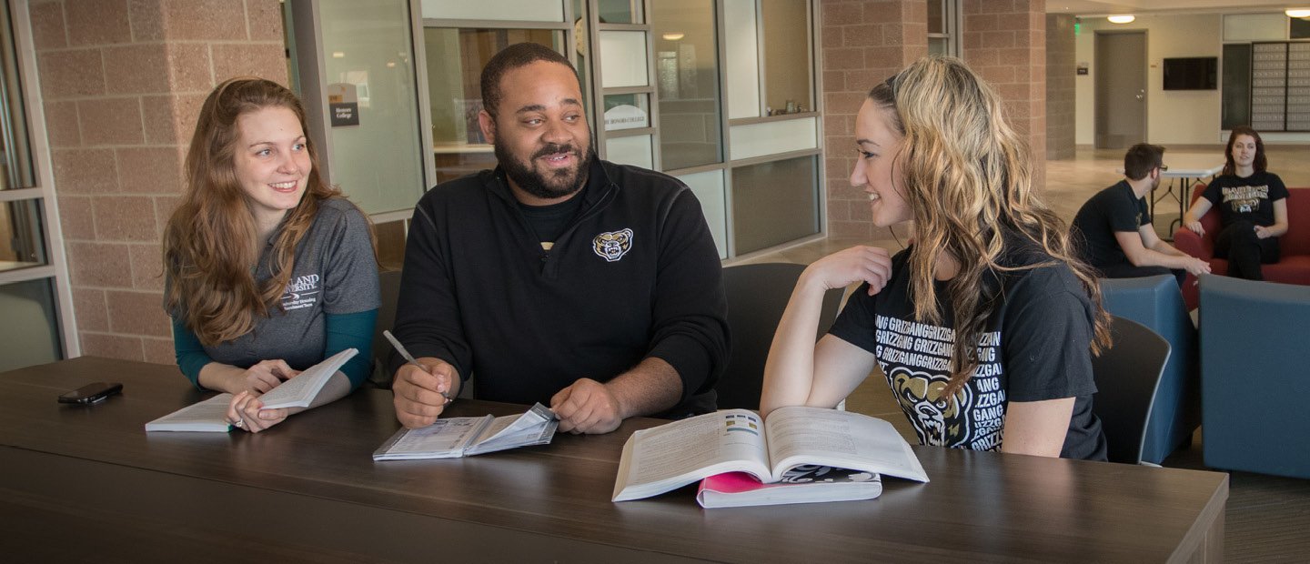 three people in Oakland University shirts seated at a table with open books