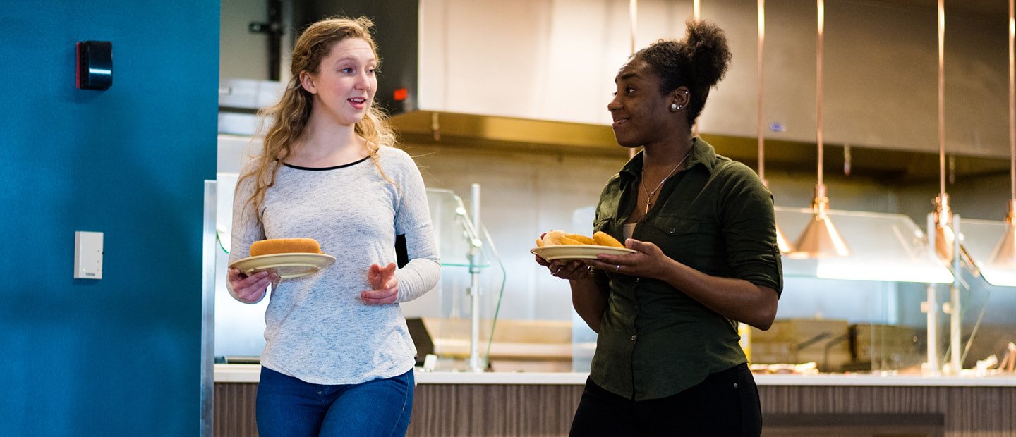 two female students carrying plates of food in a dining hall