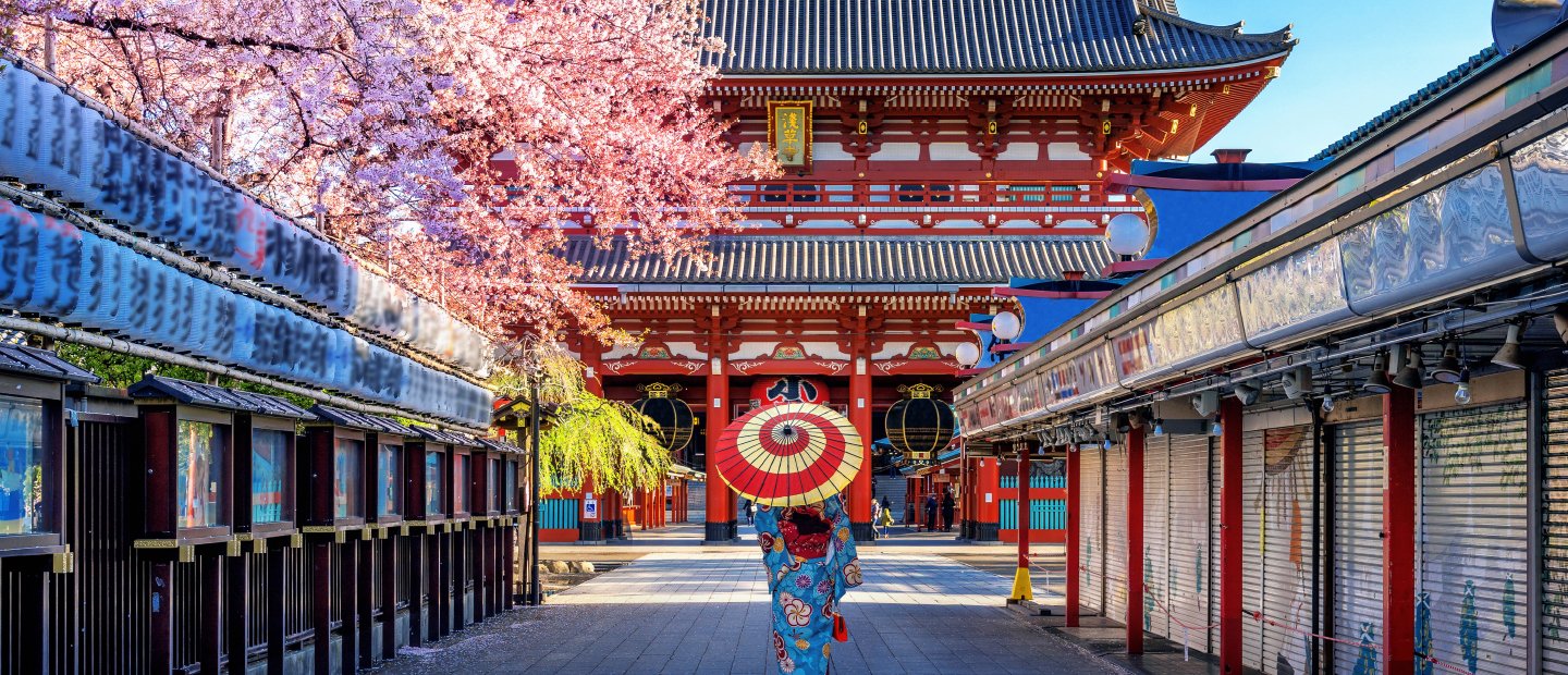 A person holding a red and white umbrella, walking on a path between buildings in Japan.