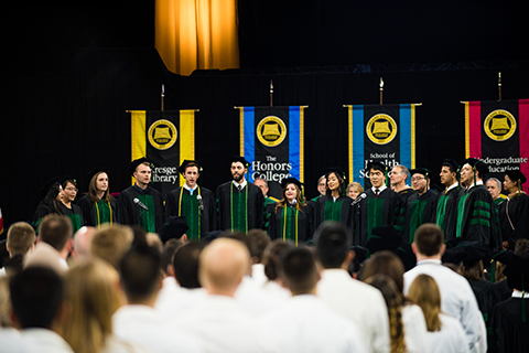 Class of 2018 members of O U W B’s DocAppella choir singing on stage in front of colored flags at their commencement