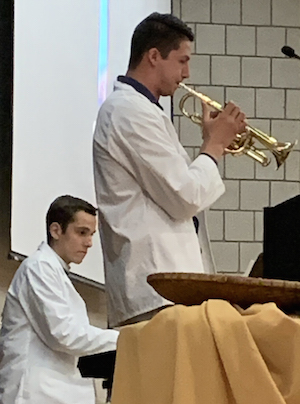 man in a white lab coat standing and playing a trumpet while another man in a white lab coat plays the piano