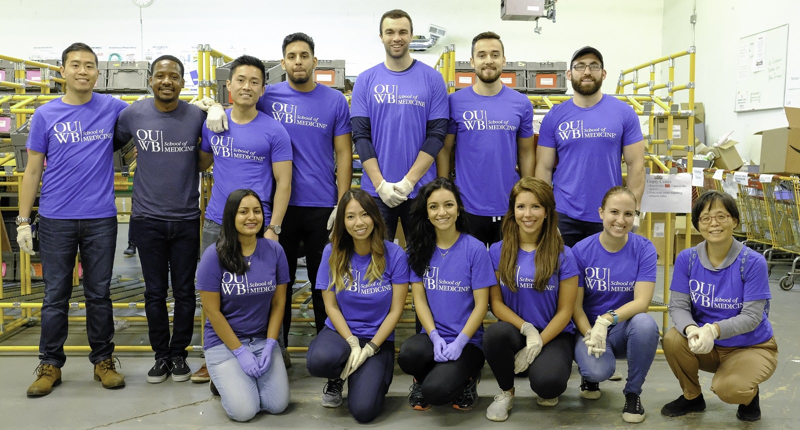 A group photo of OUWB students volunteering at World Medical Relief. The group is wearing their blue OUWB t-shirts and standing in front of a large shelf of medical supplies. 