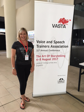 Lynnae Lehfeldt in Singapore at the V A S T A conference, standing next to a banner that says Voice and Speech Trainers Association 31st Annual Conference