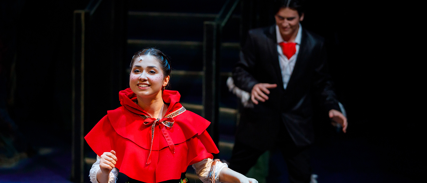 image of a theatre performance, with a girl smiling in forefront and a young man sneaking up behind her
