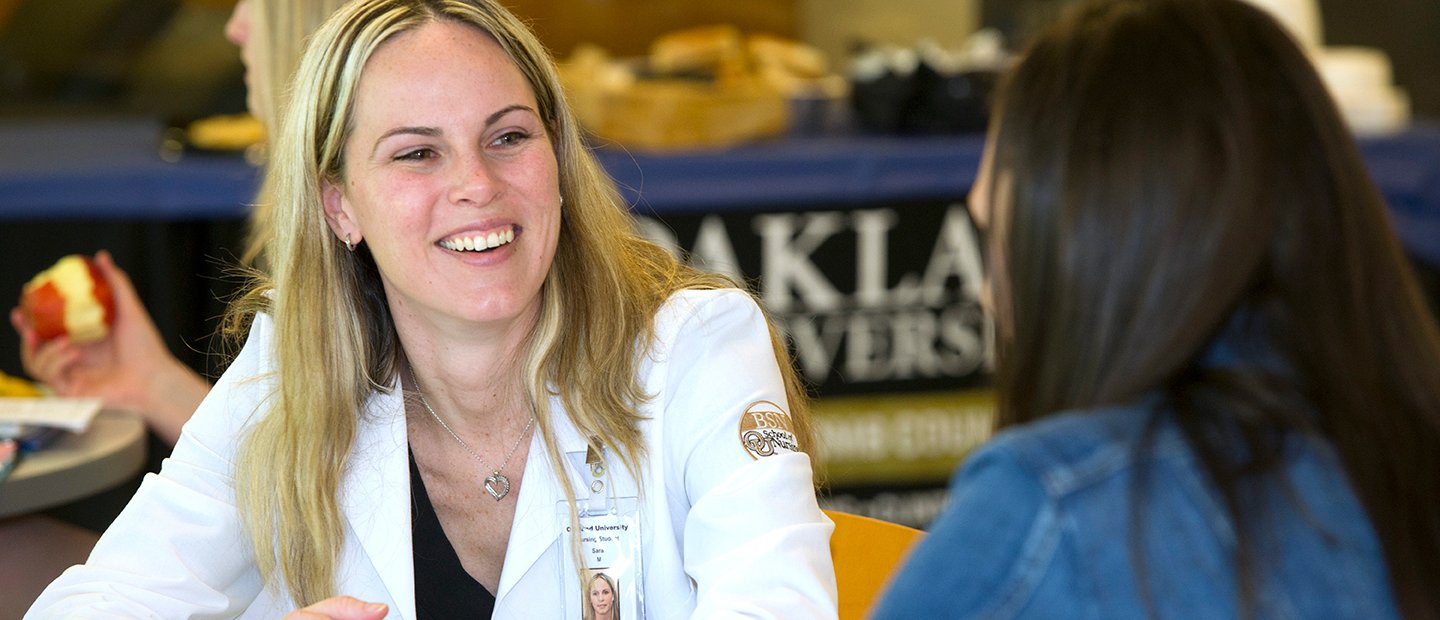 woman in a white lab coat smiling, speaking to another woman in front of her