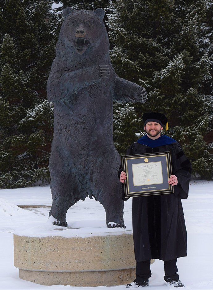 A man posing by a bear stature holding his diploma