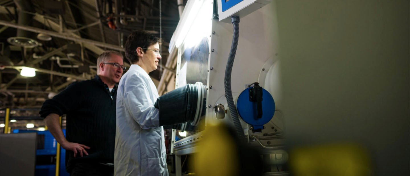 A woman in a white lab coat working at a large machine while a man watches over her shoulder.