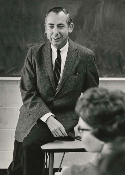 Oakland University Professor Emeritus William Schwab circa 1966, leaning on a desk speaking to a class of students with a chalkboard in the background.