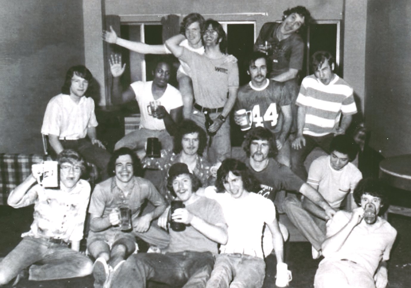 black and white photo of group of college friends from the 1970s sit and stand in a group smiling for the camera