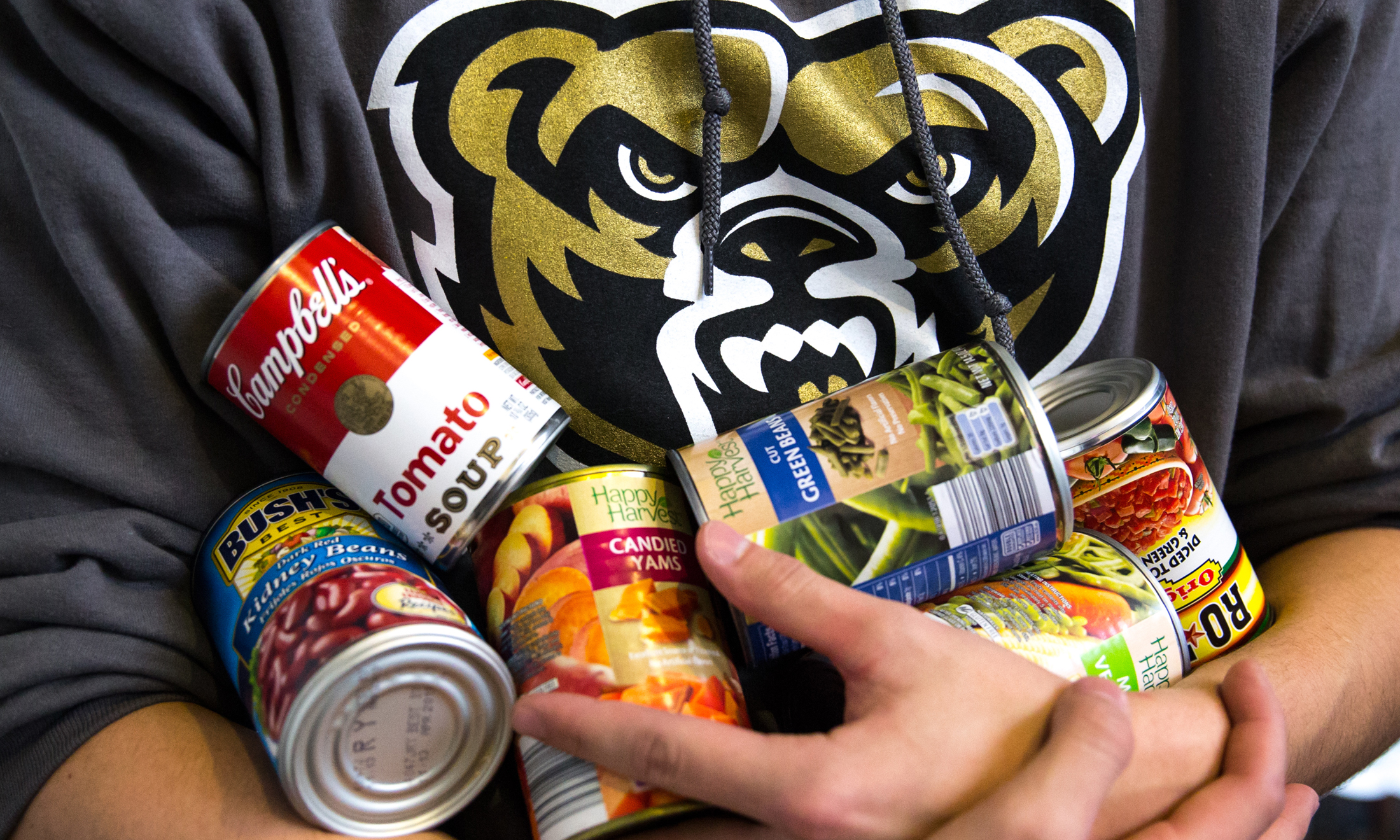 Oakland University student in gray sweatshirt with a golden grizzly head on the front hold cans of food at food drive