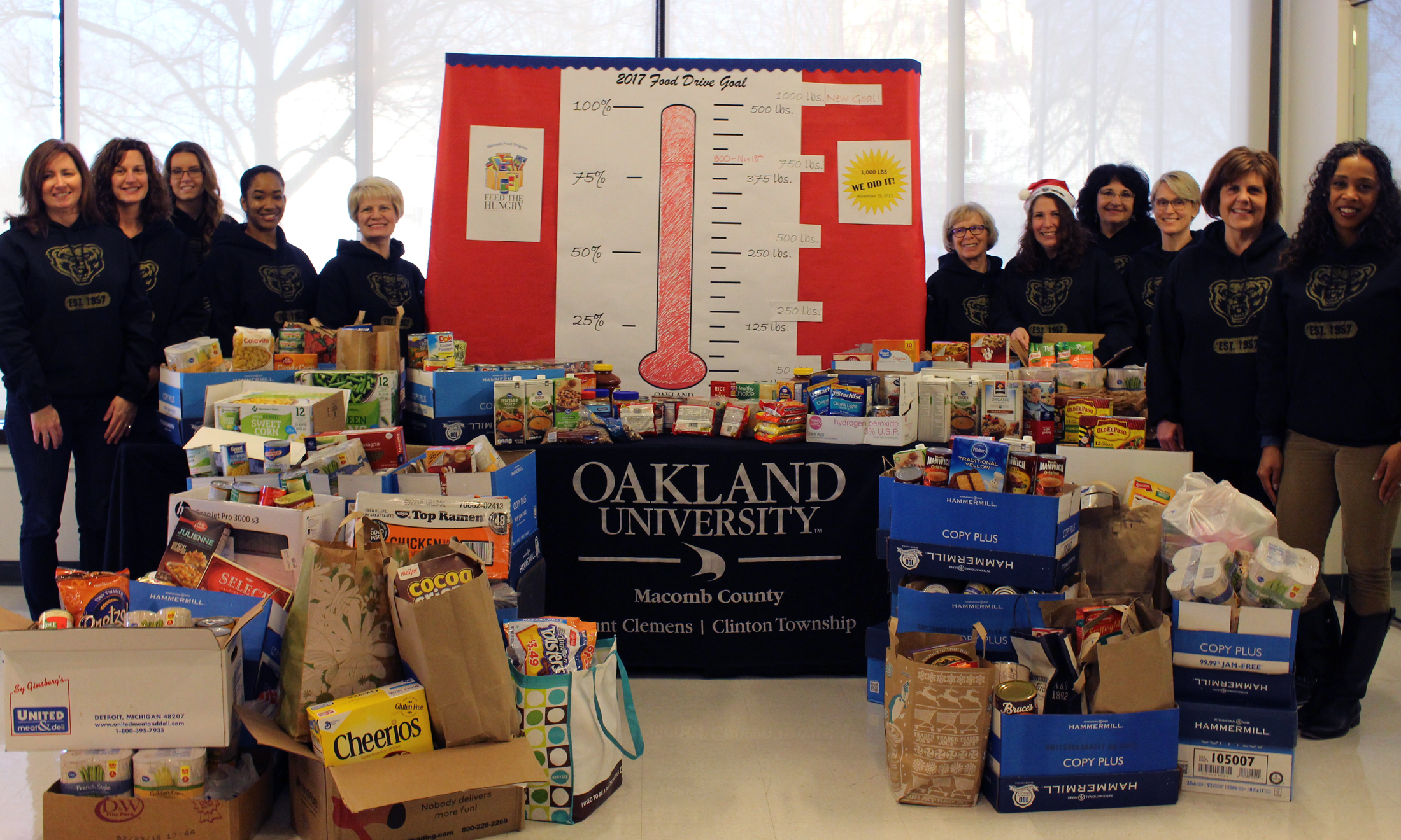 Oakland University faculty, students and staff in black golden grizzly sweatshirts stand behind a table overflowing with canned food in celebration of exceeding their food drive goal in Macomb County