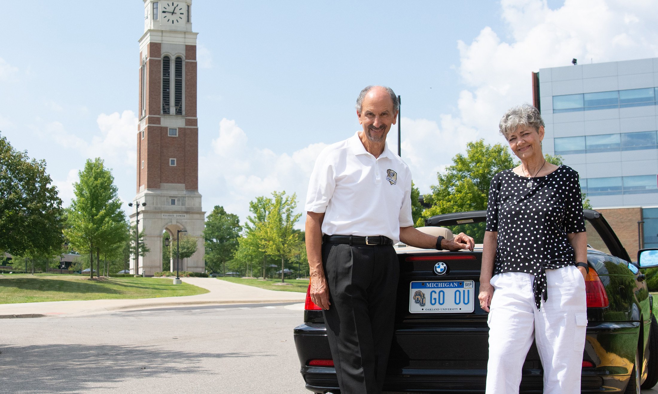 A man and woman posing in front of the rear-end of a car with a license plate that reads "GO OU"
