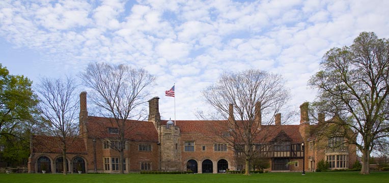 A wide shot photo of meadow brook hall in the Spring time. Blue sky with some clouds and budding trees.