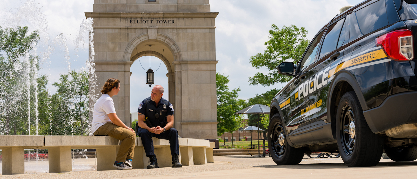 A police officer sitting with a person on a bench in front of Elliott Tower.