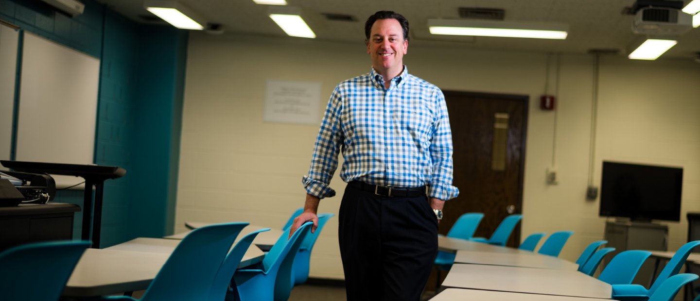 An Oakland University professor smiling at the camera, with his hand on a chair in a classroom.