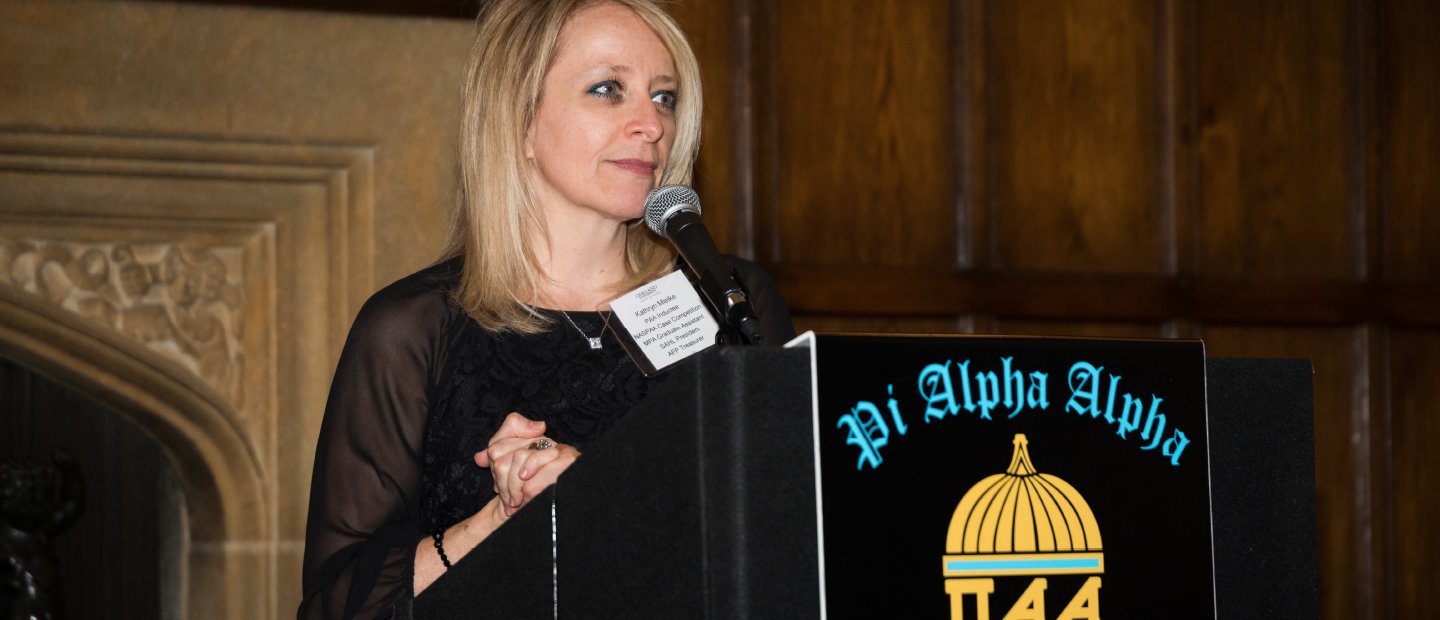 A woman standing at a podium with the words Pi Alpha Alpha on it and a graphic image of a domed capitol building.