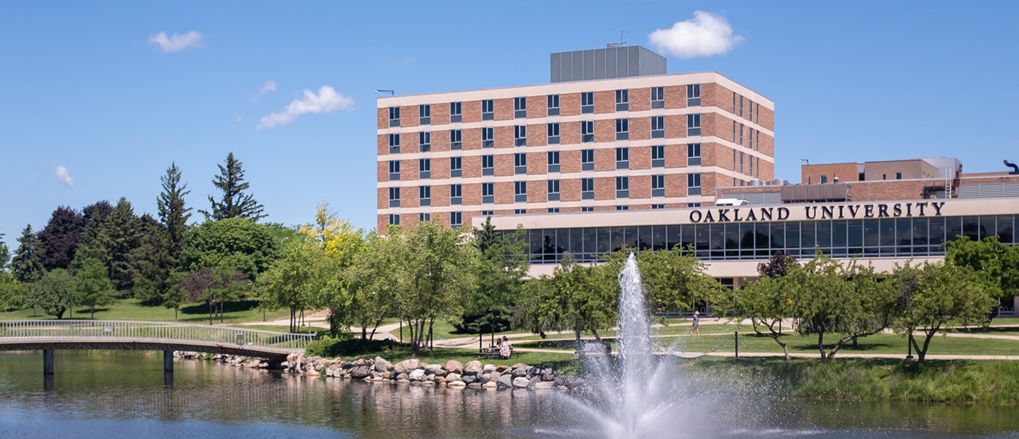 lake with a fountain, beige and brown building with Oakland University on it in the background