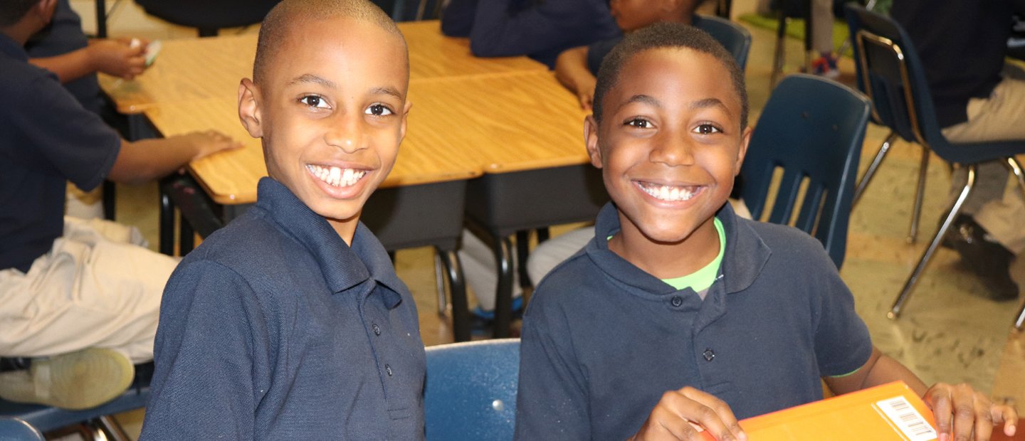 Two young boys seated in a classroom, smiling at the camera.