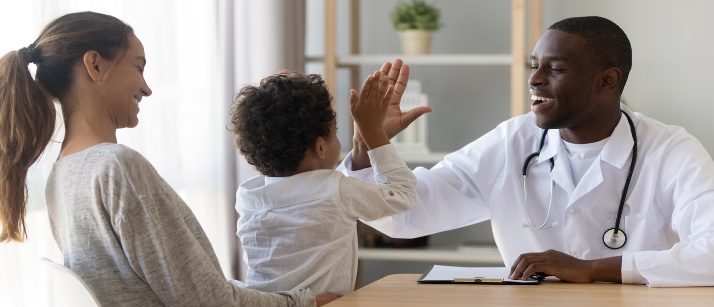 A physician's assistant giving a high five to a kid sitting in his mom's lap.