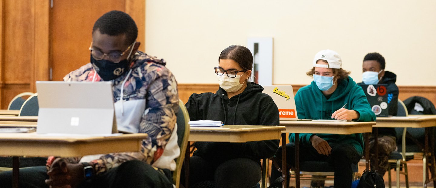 Four students sitting at individual desks in a row, all wearing face masks.