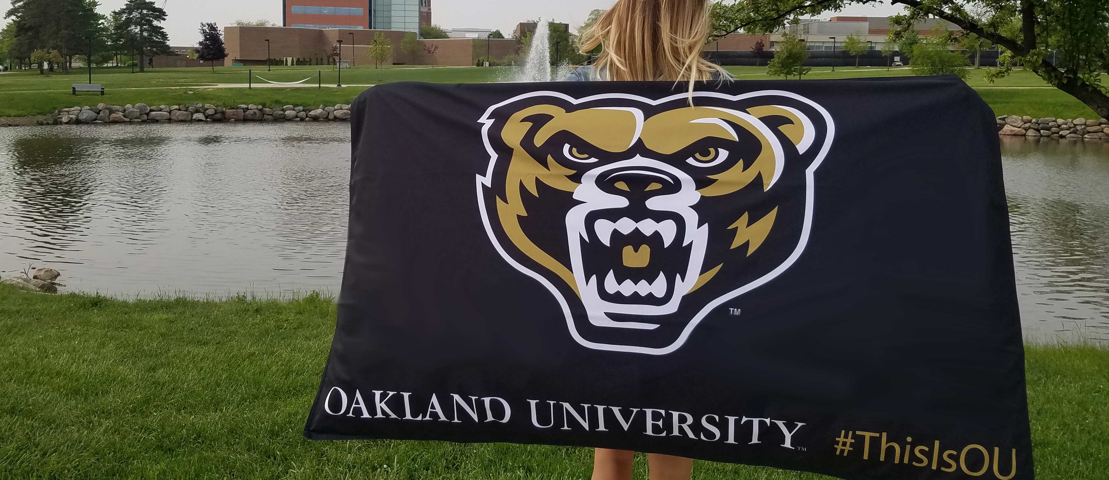 Woman holding a 3-foot by 5-foot flag in front of Bear Lake. Flag has the bear head and Oakland University #ThisIsOU printed on it. 