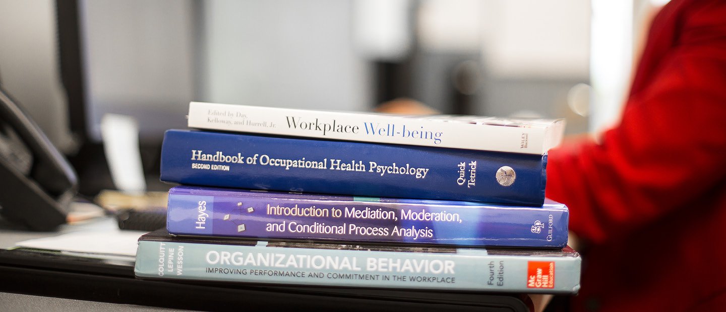 A stack of textbooks on the edge of a desk occupied by a person wearing red.